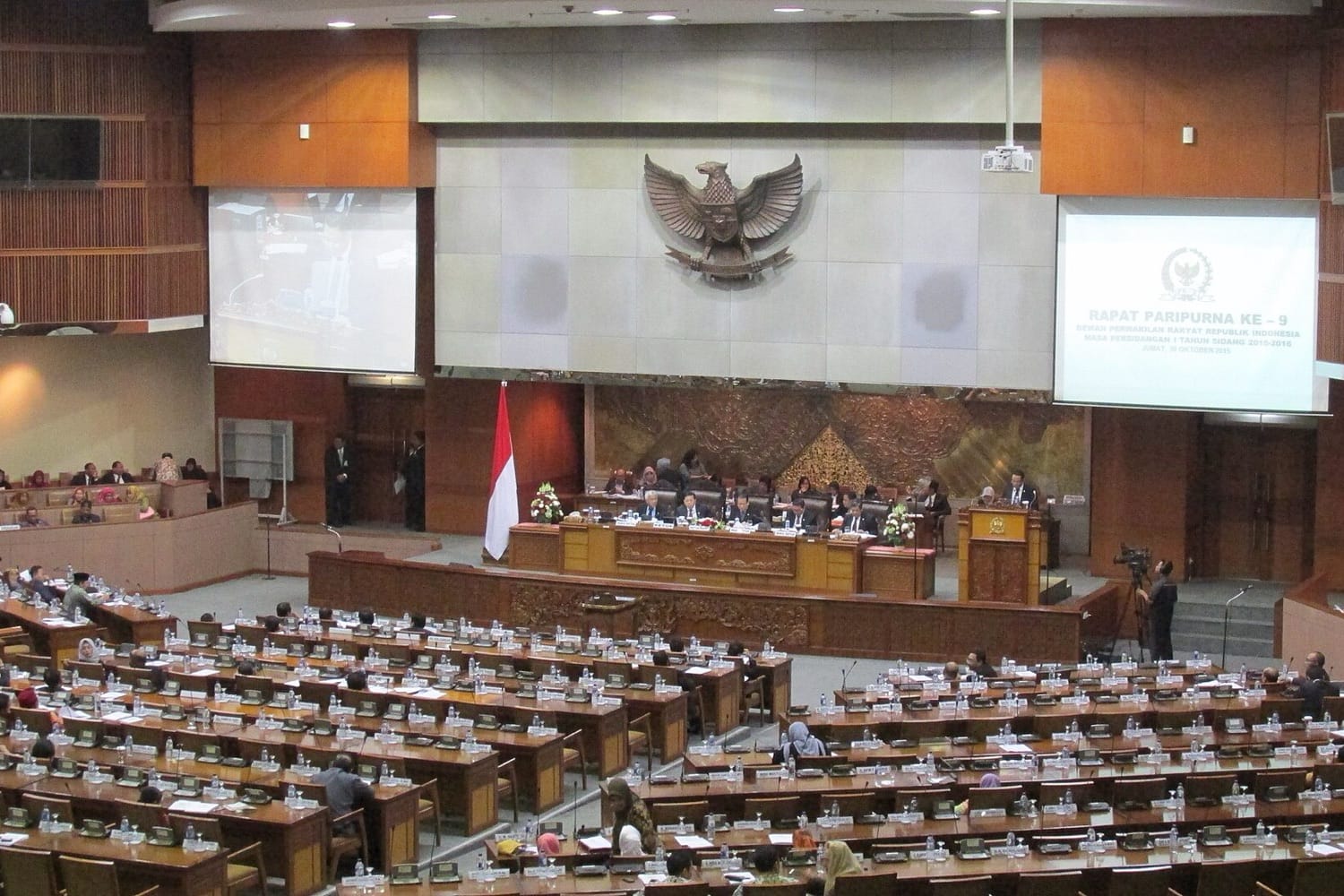 Ruang sidang paripurna DPR dengan lambang Garuda di dinding belakang dan deretan kursi anggota